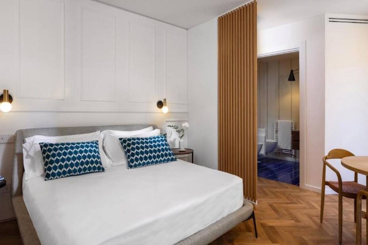 Habitación de hotel con cama, mesitas de noche, un escritorio con silla, y una zona de comedor con mesa y sillas.