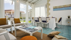 Een ruime kamer met een groot raam, een witte bank, een salontafel met tijdschriften, en een eethoek met stoelen en een tafel.