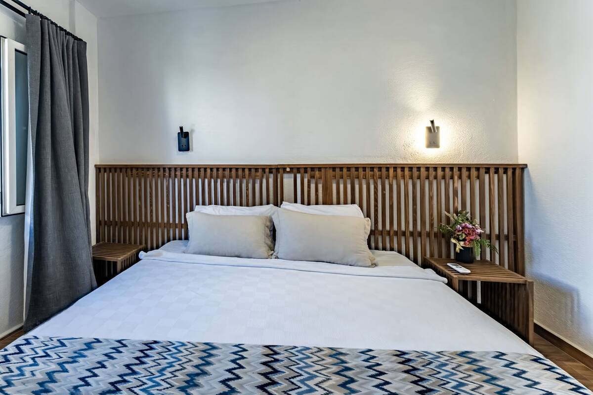 Habitación de hotel con una cama grande, cabecera de madera, dos mesitas de noche y un buró con un jarrón de flores.