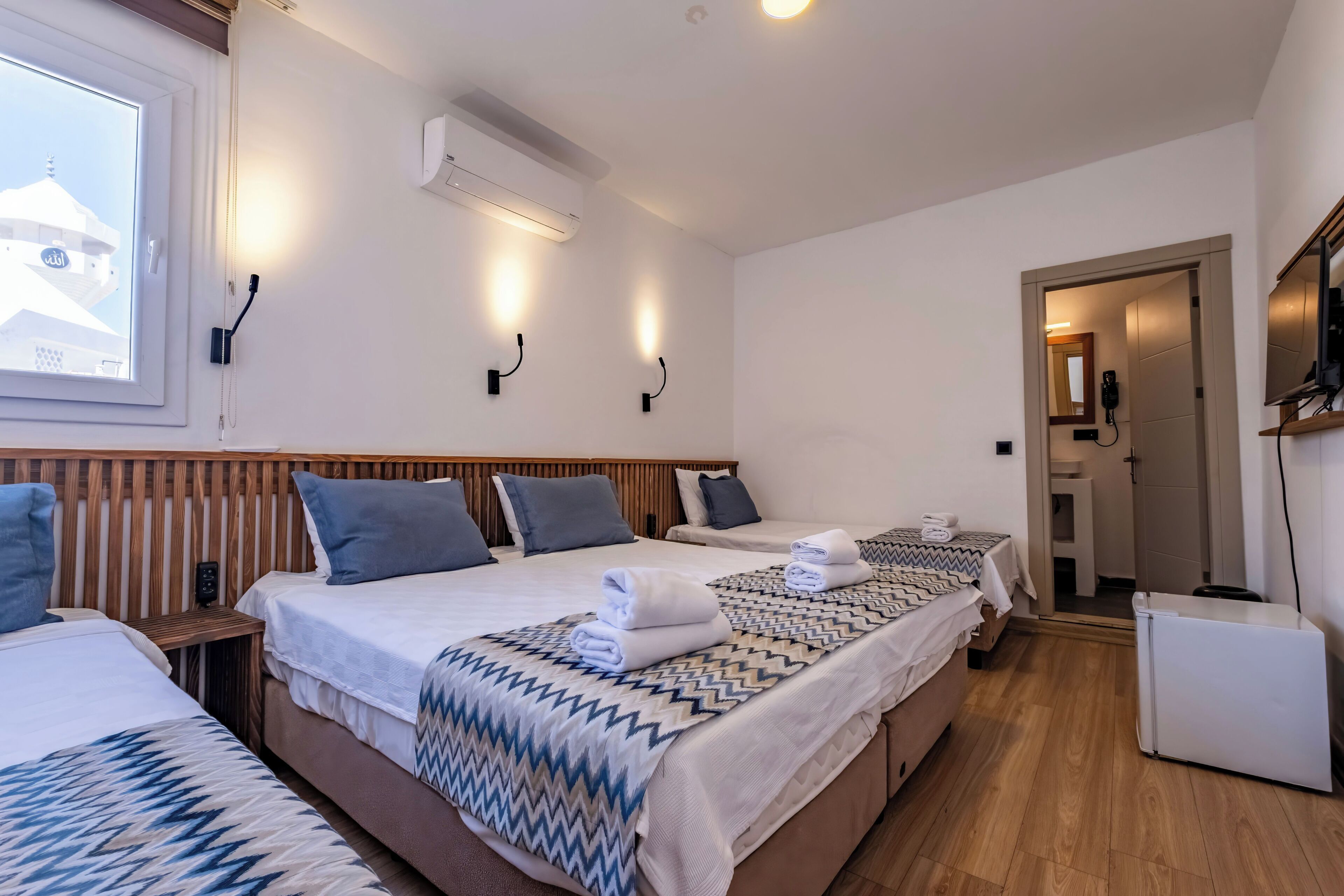 A hotel room with two beds, a TV, a refrigerator, and a window with a view of a building.