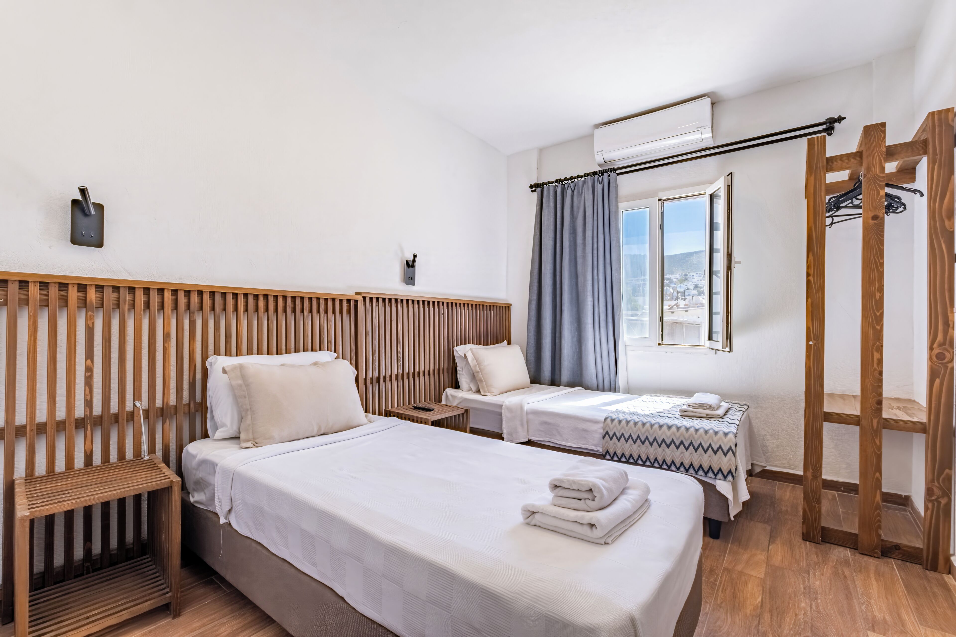Habitación de hotel con dos camas, cabecera de madera, ventana con cortinas, perchero y aire acondicionado instalado en la pared.