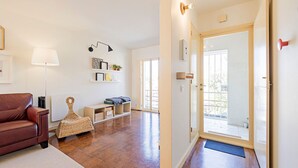 Un amplio salón con un sofá de cuero marrón, un pouf tejido y una pequeña mesa de madera. Una pared blanca con una lámpara negra y una repisa con objetos decorativos. Una puerta da a un balcón exterior.