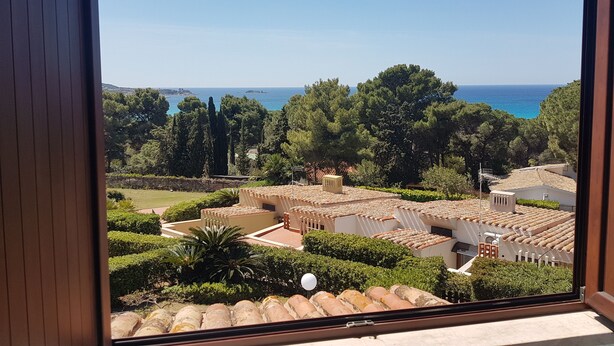 A view from a window overlooking a courtyard with a pool, surrounded by trees and buildings with Mediterranean architecture. - Apartment in a residential complex 5 minutes from Campulongu Beach (Villasimius)