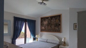 A bedroom with a bed, two bedside tables with lamps, a ceiling fan, a large framed picture on the wall, and a window with blue curtains. - Apartment in a residential complex 5 minutes from Campulongu Beach (Villasimius)