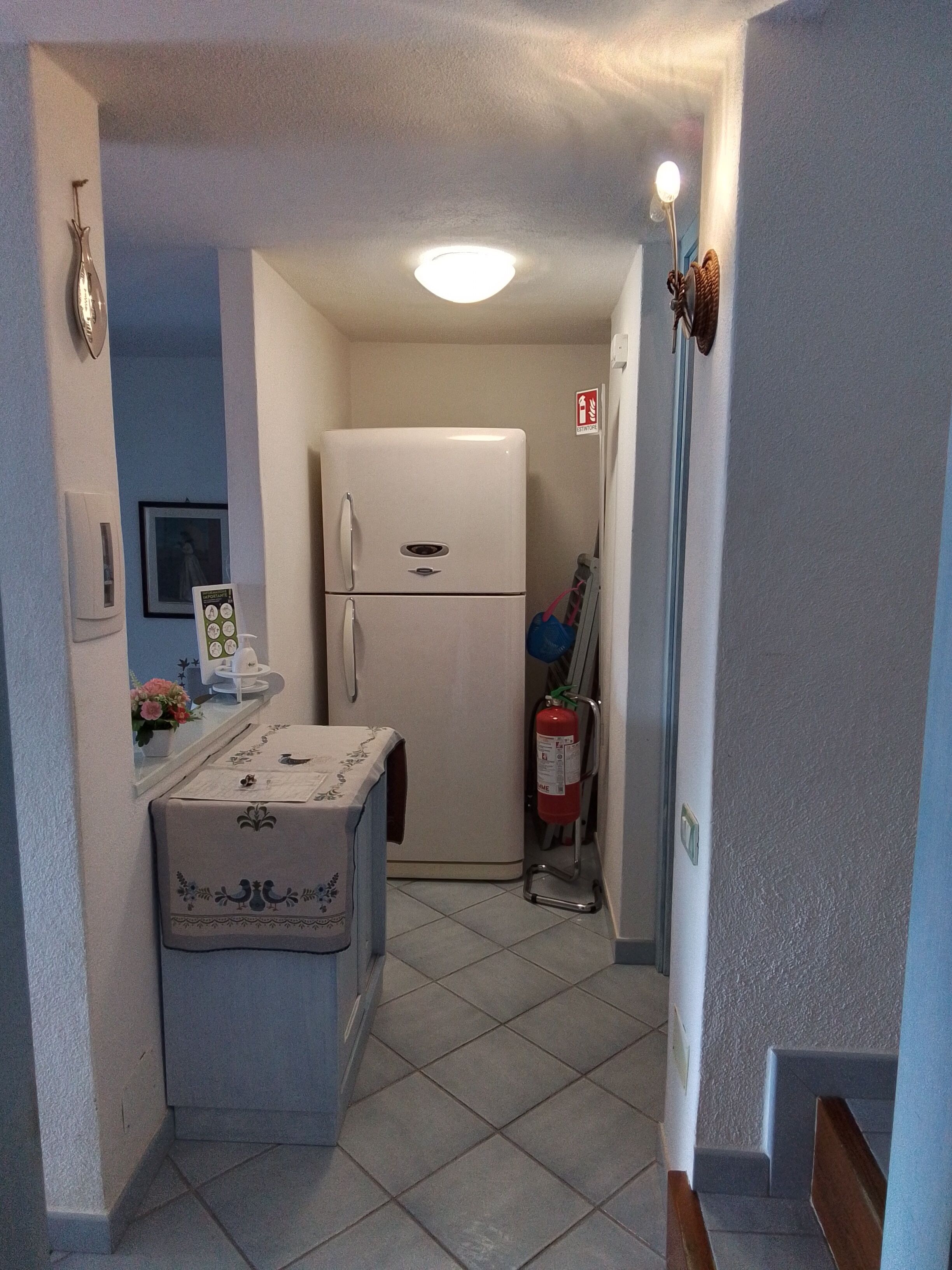 A narrow hallway with a white refrigerator, a small table with a floral tablecloth, and a fire extinguisher.