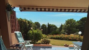 An outdoor patio area with tiled flooring, a covered seating area with chairs and a table, and a view of a garden with various plants and a lawn. - Apartment in a residential complex 5 minutes from Campulongu Beach (Villasimius)