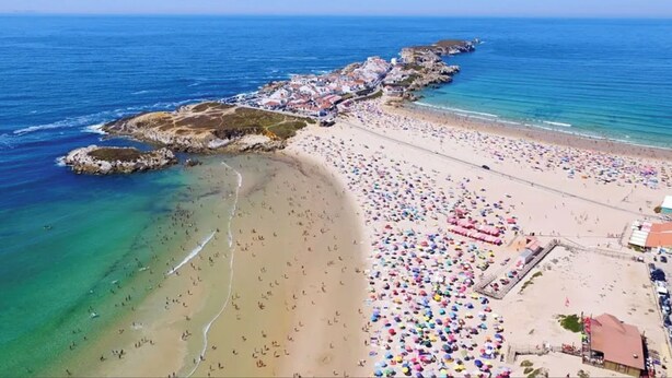 Vista aerea di una spiaggia con costa rocciosa, zona di spiaggia sabbiosa e un gruppo di ombrelloni colorati.