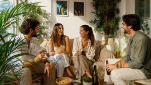 Un gruppo di persone sedute su un divano in una stanza con ampie finestre, una pianta in vaso e un tavolo con una bottiglia e una ciotola.