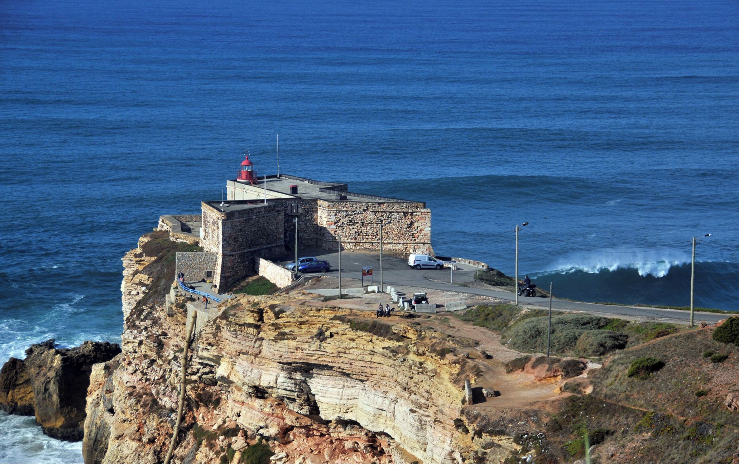 A coastal cliff with a lighthouse, a paved area with cars, and a road leading to the sea.