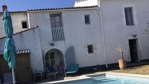 A swimming pool with a wooden deck, a covered patio area with chairs, and a white building with a tiled roof. - Ile de re family house, garden swimming pool, close to the sea on foot. (Sainte-Marie-de-Ré)