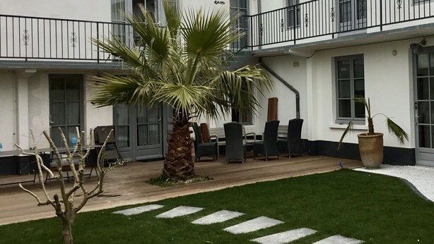 A courtyard with a palm tree, outdoor seating, and a white building with balconies. - Ile de re family house, garden swimming pool, close to the sea on foot. (Sainte-Marie-de-Ré)