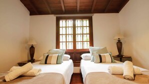 Una habitación con dos camas, cada una con una almohada y una toalla enrollada. Hay dos mesitas de noche con lámparas. Una ventana grande con múltiples divisiones se encuentra en el centro.
