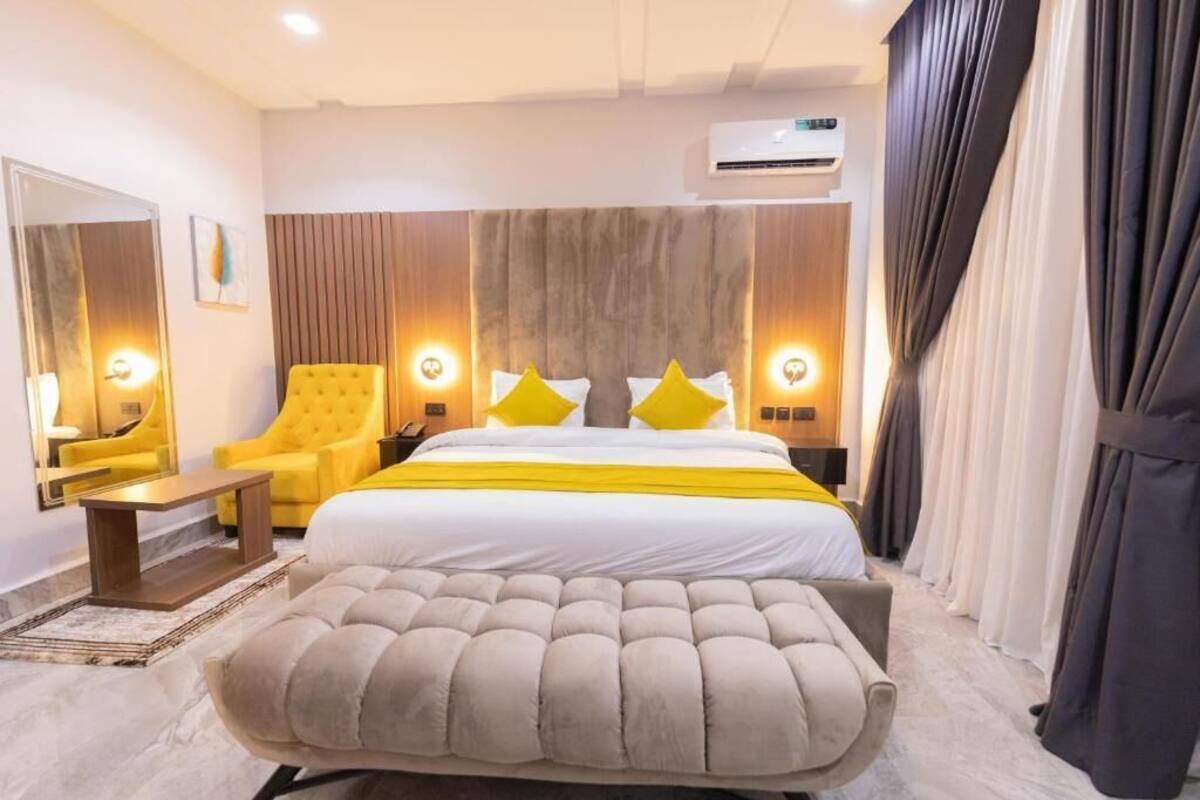 A modern hotel room with a large bed, a tufted ottoman, a yellow armchair, and a nightstand with a lamp.