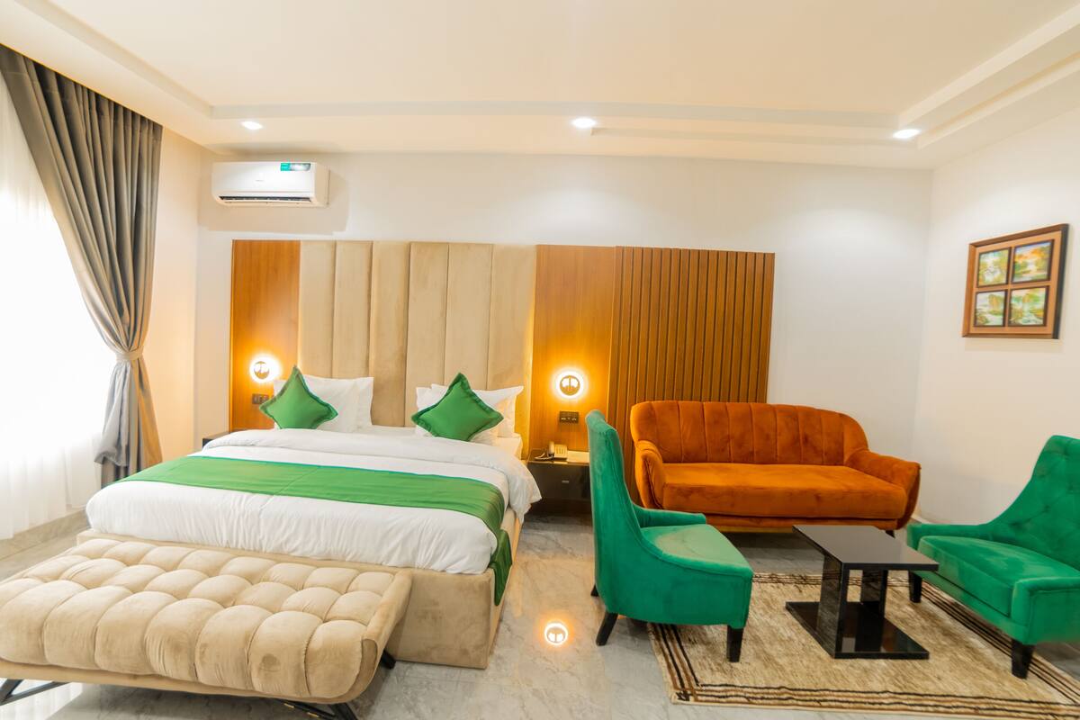 A hotel room with a bed, a sofa, two armchairs, a small table, an air conditioning unit, and a painting on the wall.