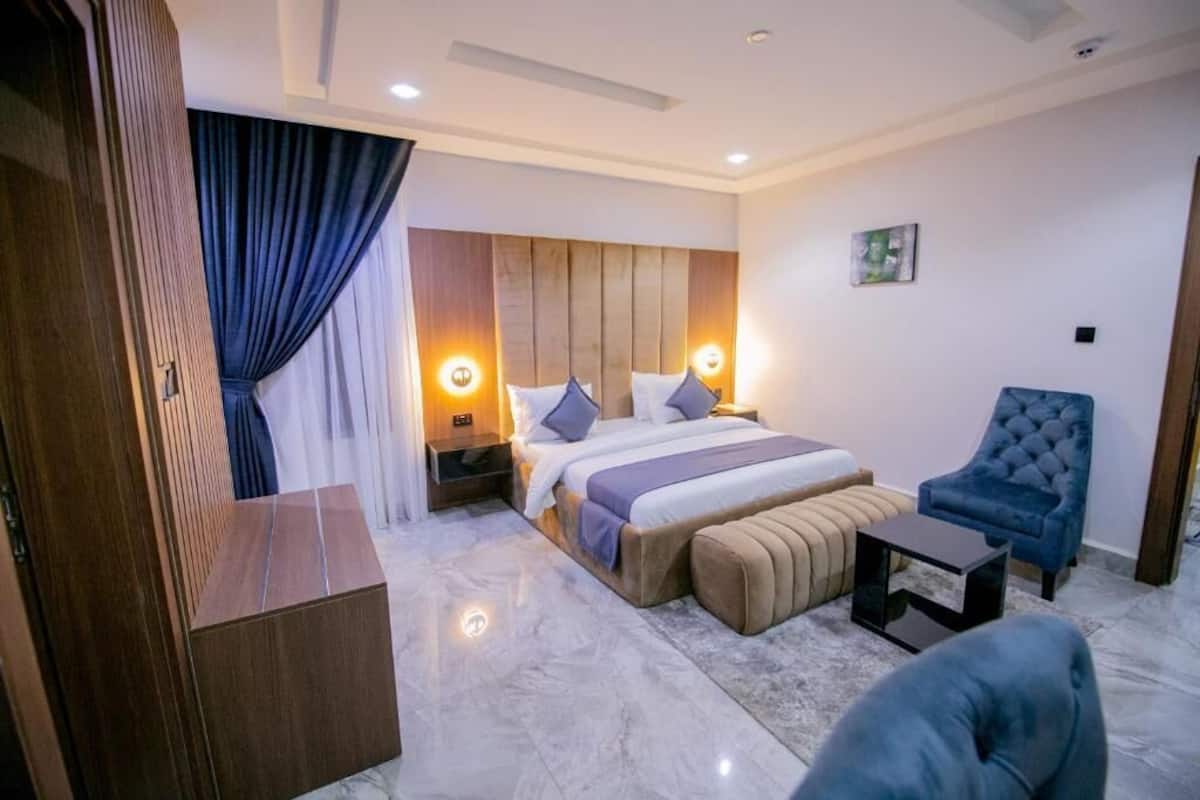 A modern hotel room with a large bed, a blue armchair, a small table, and a wooden wardrobe.