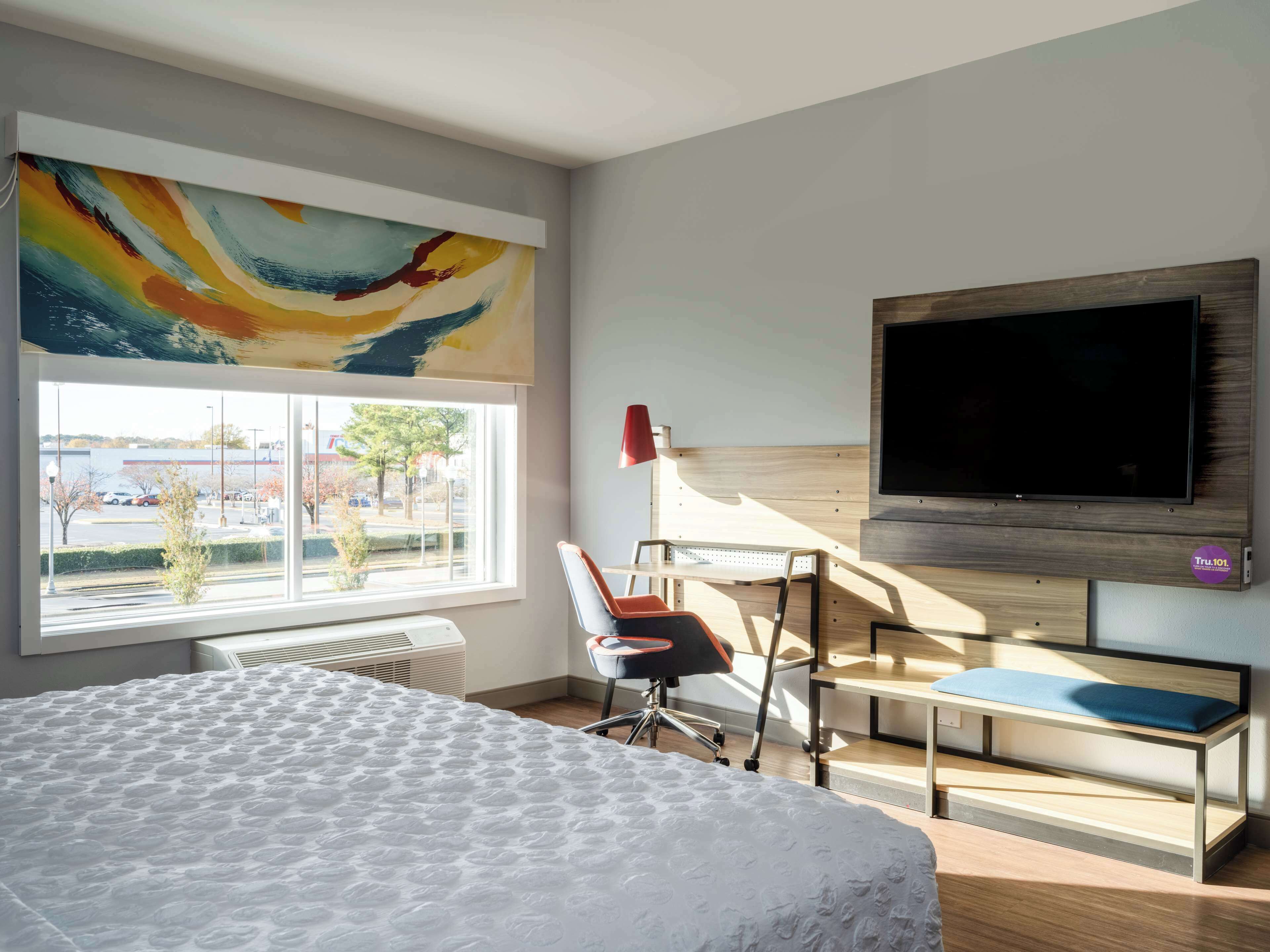 A modern hotel room with a large bed, a desk, a chair, a TV, and a window with a view of a parking lot and trees.