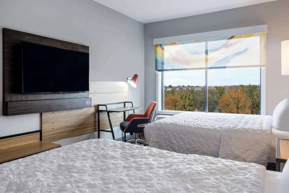 Une chambre d’hôtel avec deux lits, un bureau, une chaise, une télévision et une grande fenêtre donnant sur des arbres.