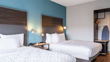 Une chambre d’hôtel avec deux lits, une tête de lit en bois, des tables de chevet avec des lampes et un petit coin équipé pour le repassage.