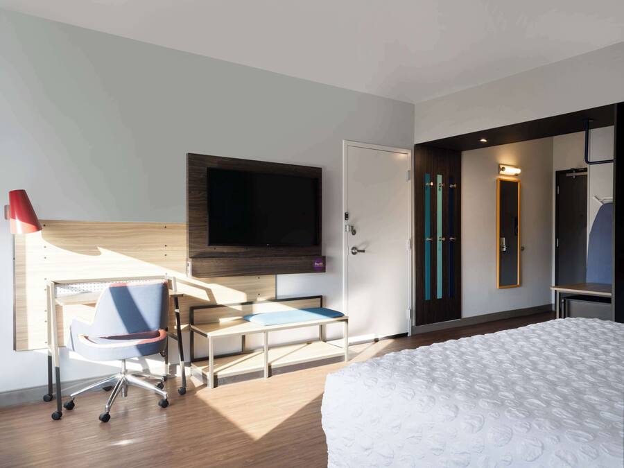 A modern hotel room with a bed, a desk with a chair, a flat-screen TV mounted on the wall, and a door leading to another room.