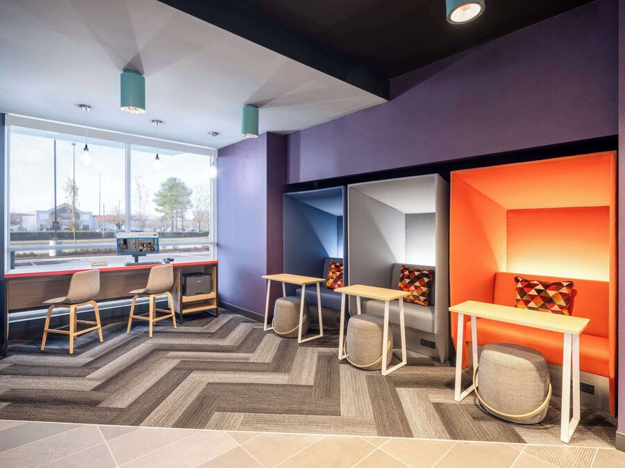 A modern office space with a herringbone-patterned carpet, multiple seating areas with colorful geometric cushions, and a large window providing a view of the outside.