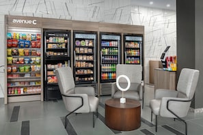 Un espacio comercial moderno con una variedad de productos de comida y bebida, como papas fritas, cereales y bebidas embotelladas. Hay dos sillas grises y una pequeña mesa redonda con una luz de aro.