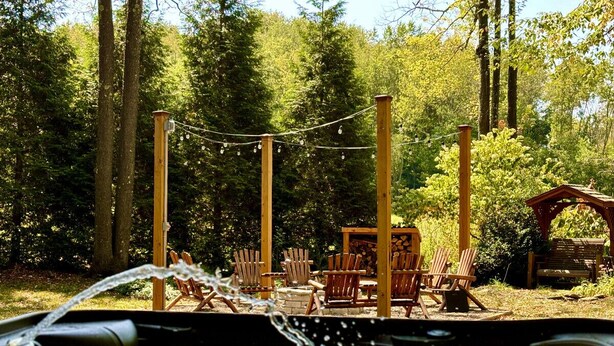Een hottub met stromend water, omringd door houten stoelen en een houten constructie in een bebost gebied.