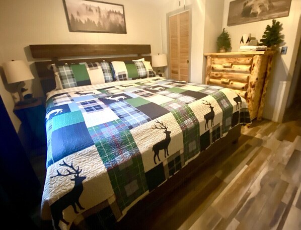 A bedroom with a large bed, a wooden headboard, a nightstand with a lamp, a dresser, and a small Christmas tree.
