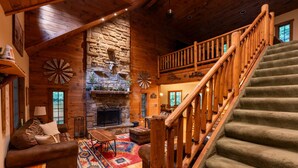 A log cabin interior with a stone fireplace, a wooden staircase, and a cozy seating area.