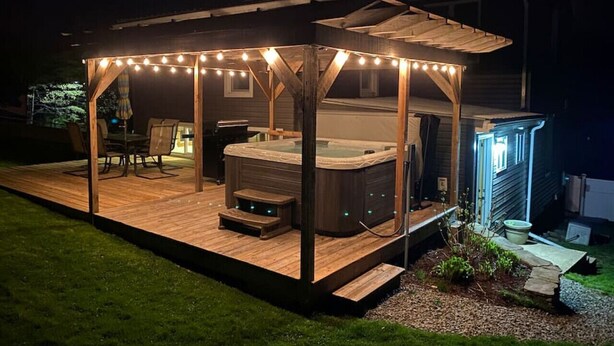 Un jacuzzi bajo una pérgola de madera con luces colgantes, rodeado por una terraza y un pequeño jardín.