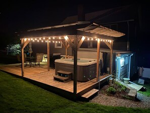 A hot tub under a wooden pergola with string lights, surrounded by a deck and a small garden area.