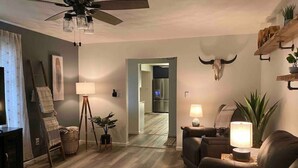 Un salon avec un ventilateur de plafond, une lampe, un canapé, un tapis et une étagère murale ornée de plantes.