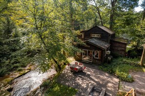 Una cabaña rústica rodeada de un bosque frondoso, con un patio y un pequeño hogar para fogata.