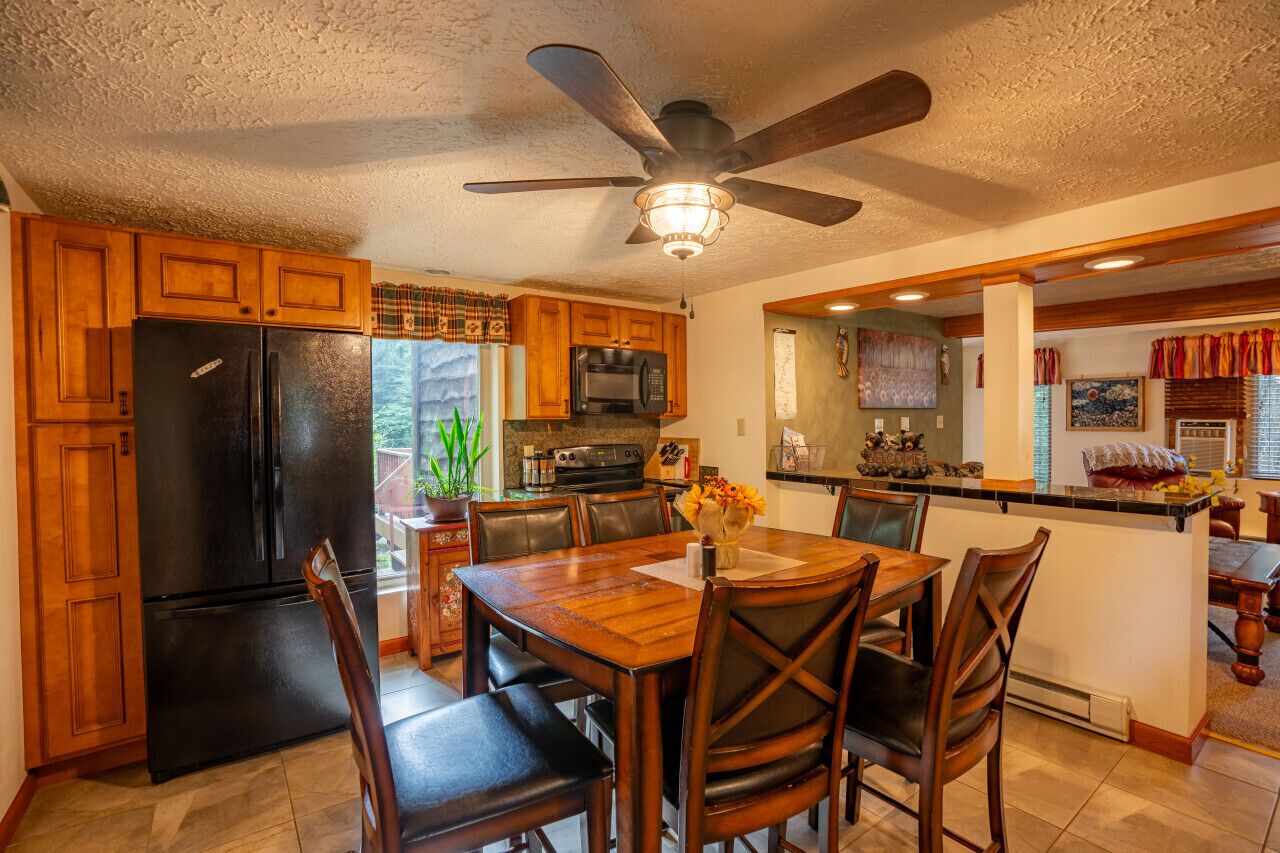 Une cuisine avec des meubles de rangement en bois, un réfrigérateur noir, une table à manger avec des chaises et un ventilateur de plafond.