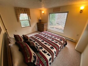 Un dormitorio con una cama grande, dos ventanas, un aparador y una lámpara de pared.