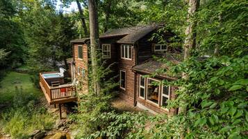 A rustic cabin with a deck and a hot tub, surrounded by dense forest.