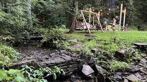 Tunnelmallinen, maalaishenkinen puinen keinuteline vehreässä metsässä, lähellä pientä puroa.