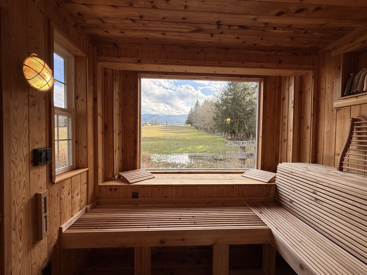 A wooden cabin interior with a bench, a window with a view of a grassy field, and a light fixture.