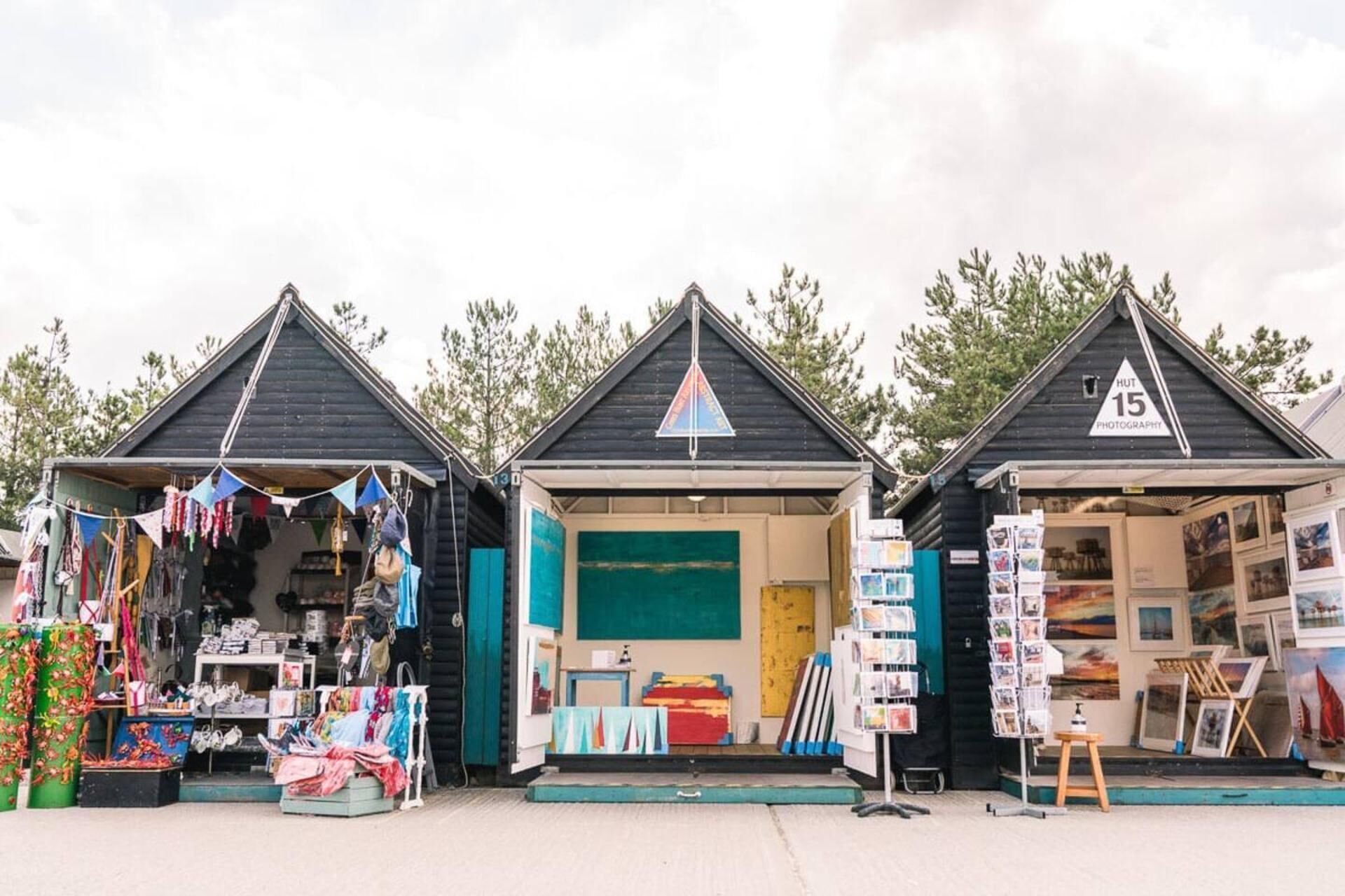 Three small, dark-colored huts with triangular roofs, each displaying different items for sale.