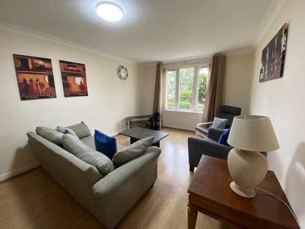 A living room with a sofa, armchairs, a coffee table, and a lamp. There are two windows with curtains, a clock on the wall, and two framed pictures. - Pure Apartments Dunfermline East - Dalgety Bay (Saint Davids)