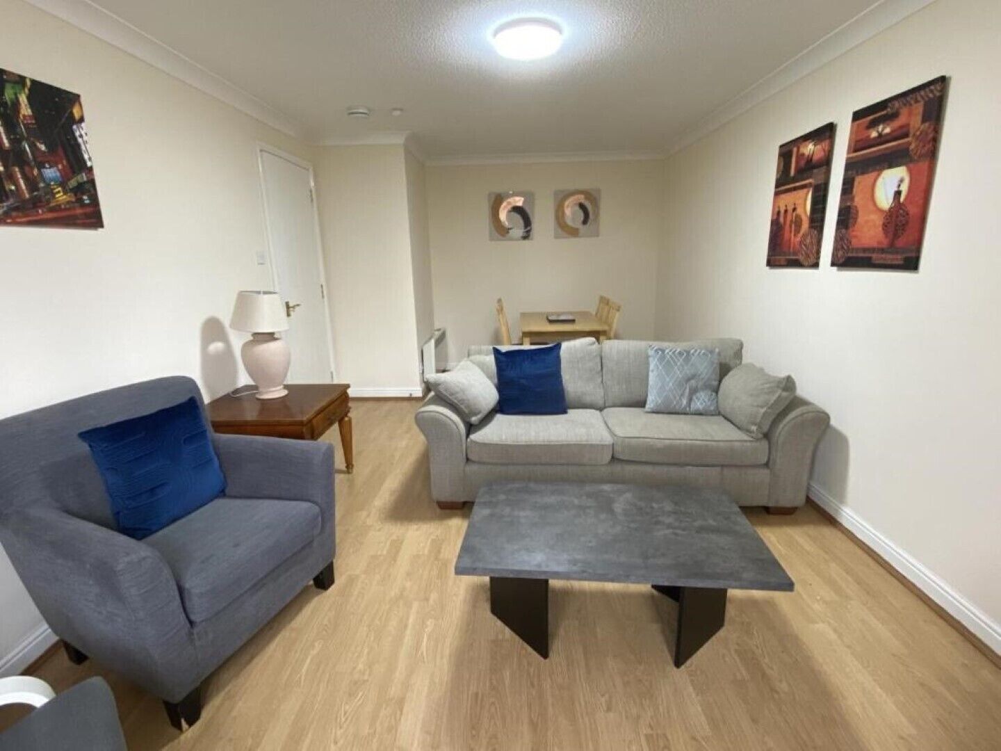 A living room with a grey sofa, a grey armchair, a grey sofa with blue cushions, a coffee table, and a wooden side table with a lamp. There are two wall-mounted paintings and a door in the background.
