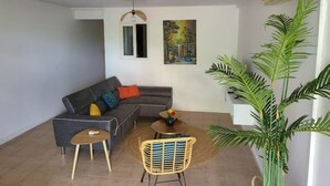 En moderne stue med en grå sofa, et lille rundt bord og en fletstol. Der er en stor stueplante og et billede på væggen.