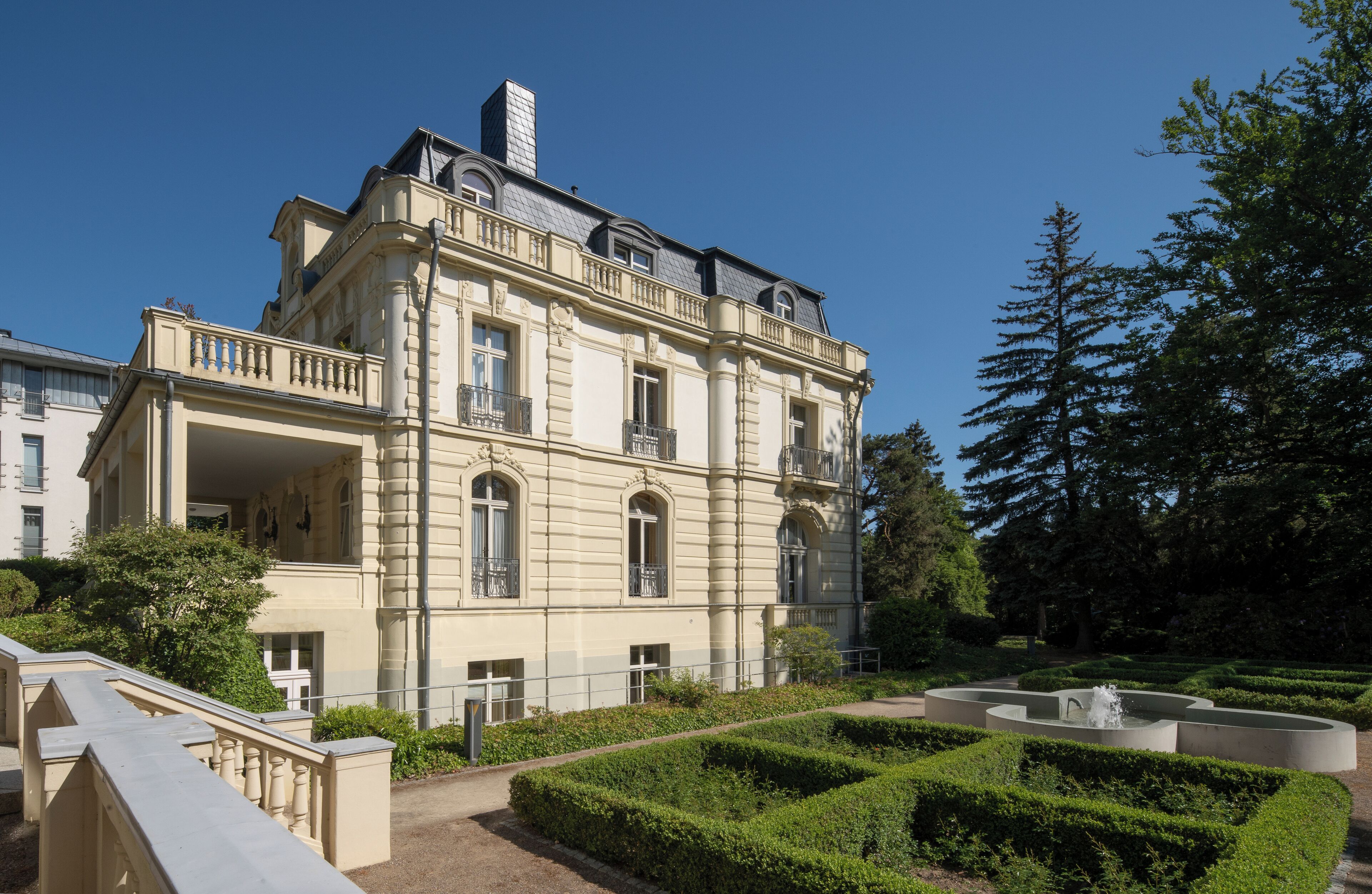 Un edificio grande de estilo clásico con múltiples balcones y un jardín formal con una fuente.