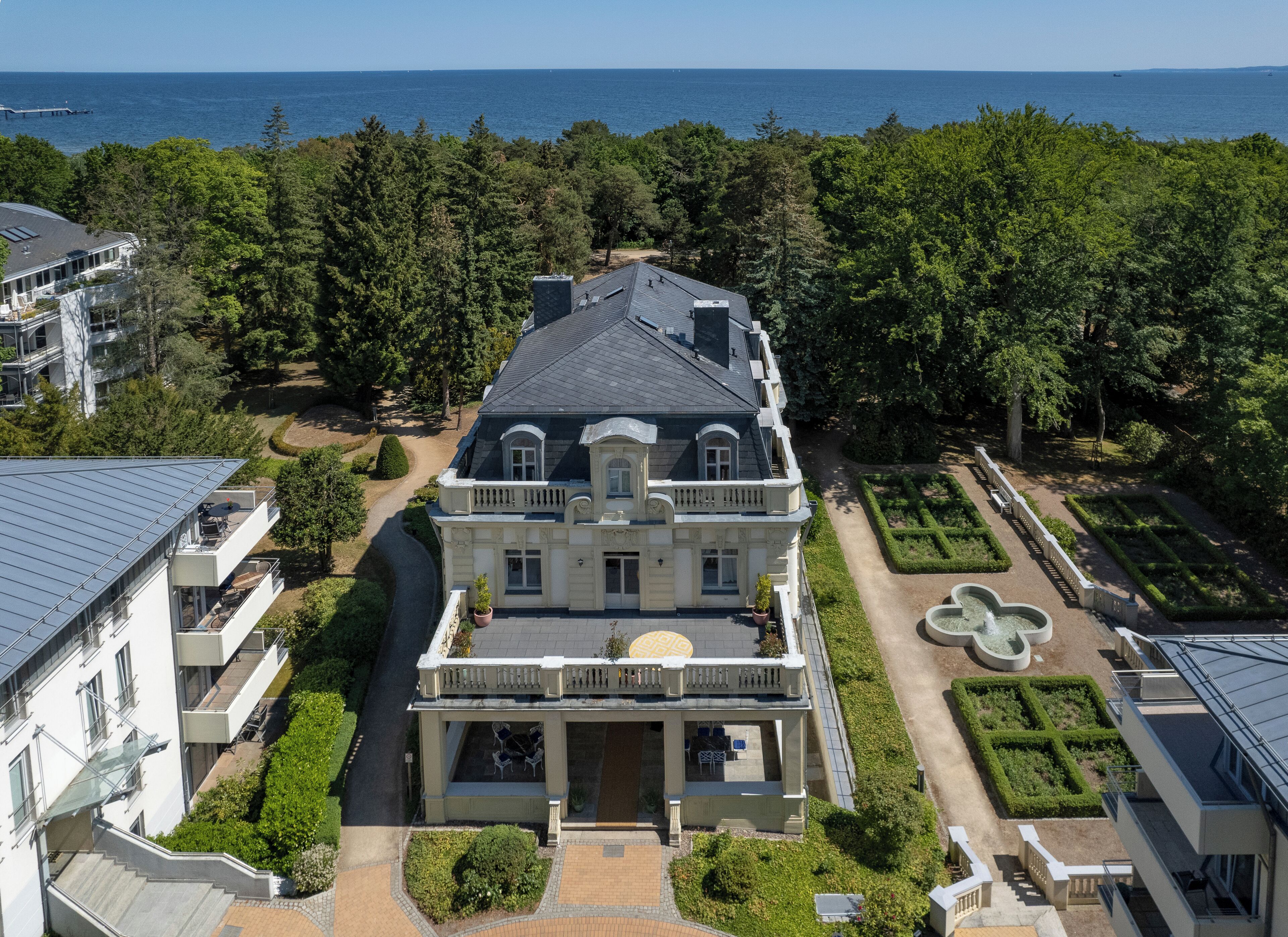 A large, historic building with a symmetrical facade, a central entrance, and a formal garden with a fountain.