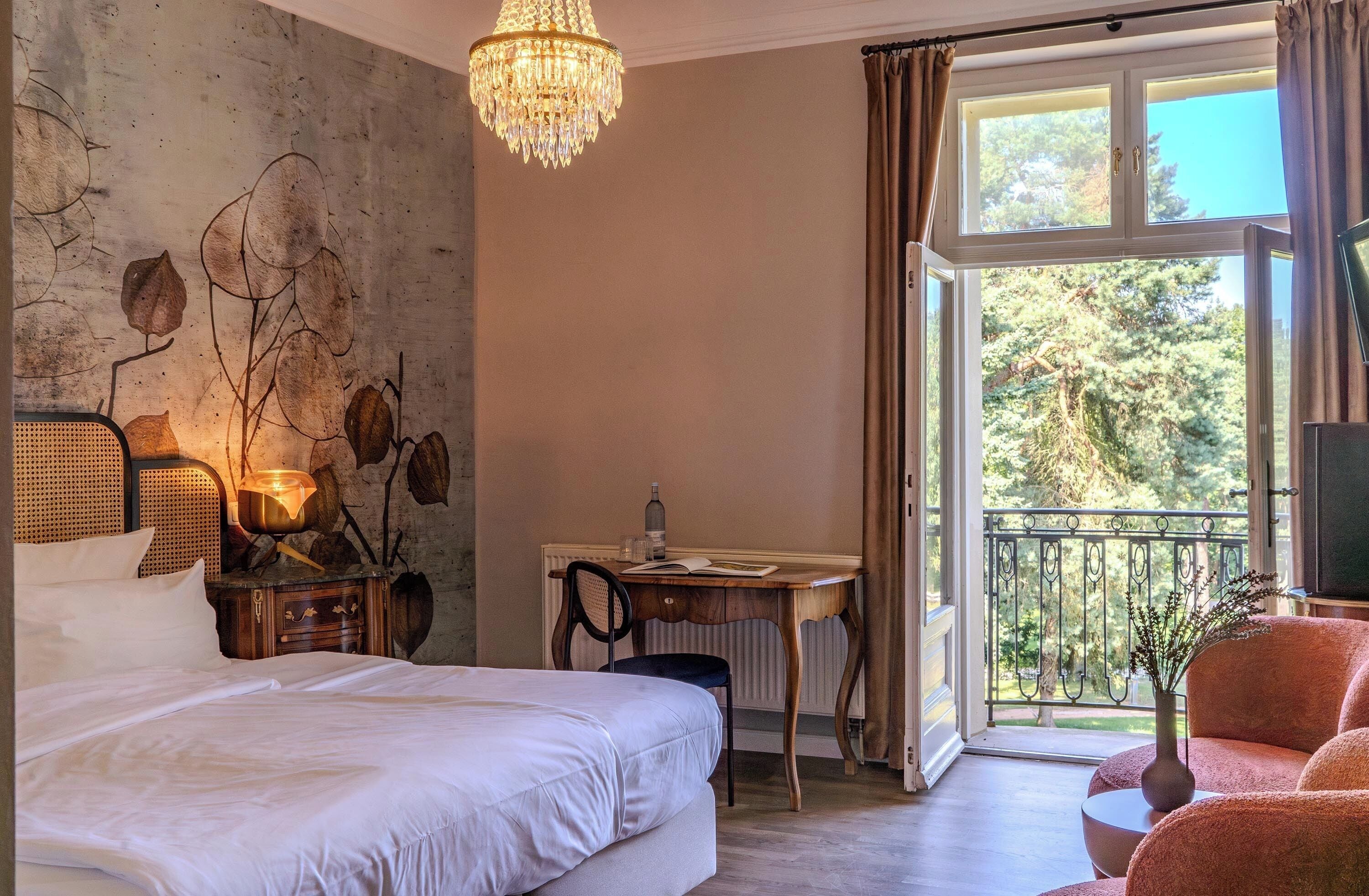 A bedroom with a bed, a desk, a chair, a chandelier, and a view of the outdoors.
