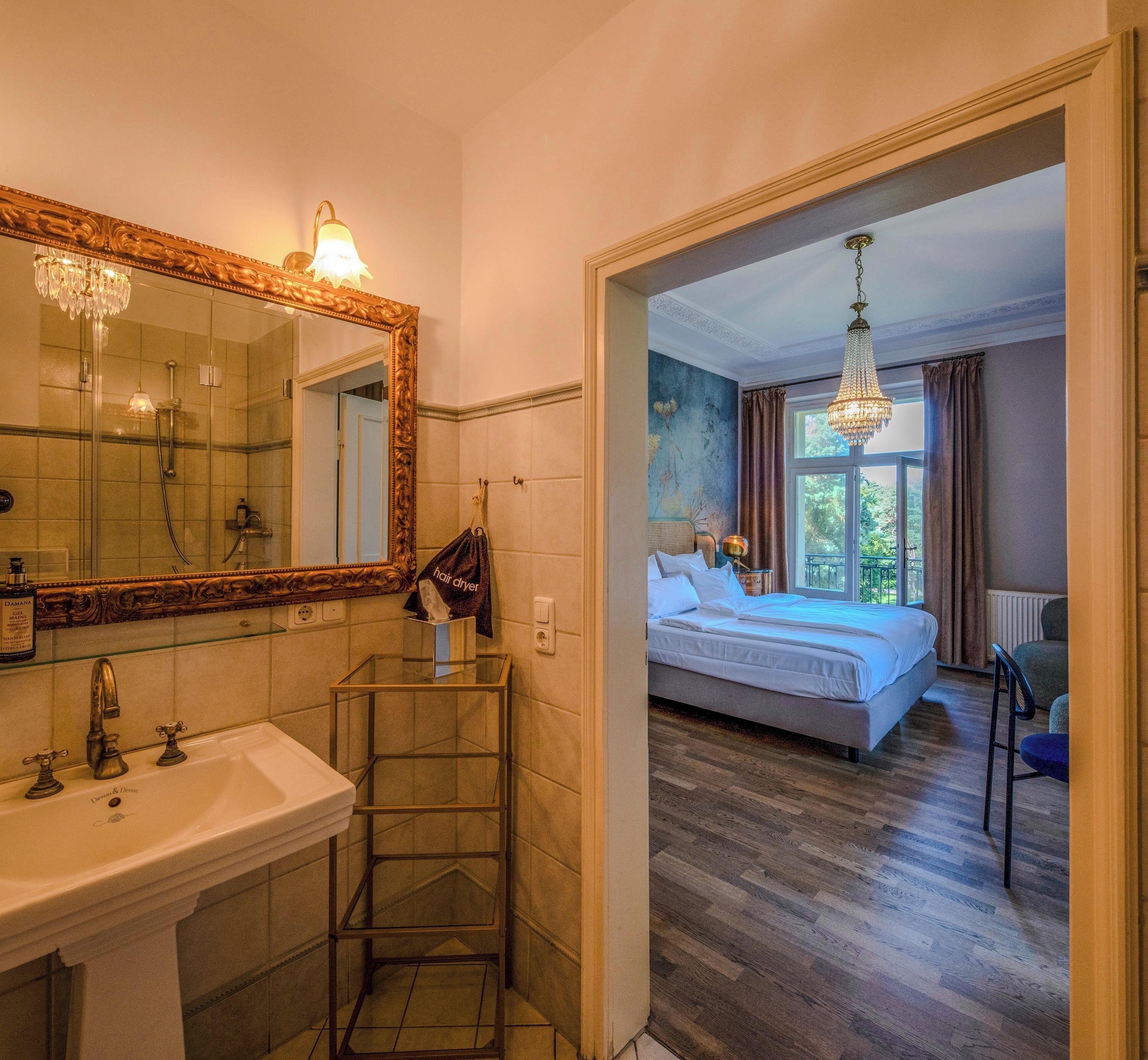 Een badkamer met een grote spiegel, wastafel en douche. De kamer heeft een houten vloer en een bed met uitzicht op groen buiten.