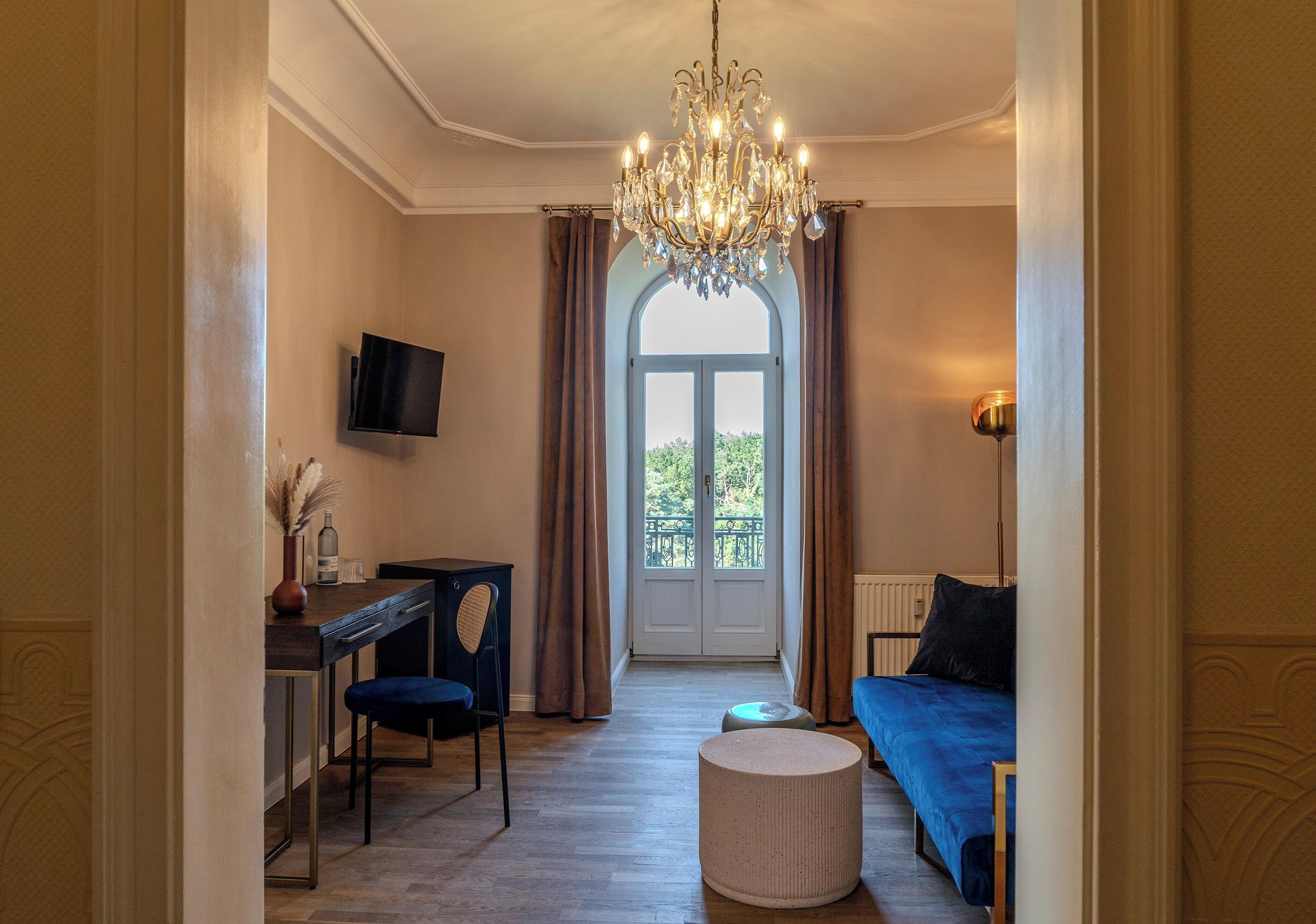 Quarto com lustre, televisão, sofá, escrivaninha com cadeira e vista através de uma grande janela.