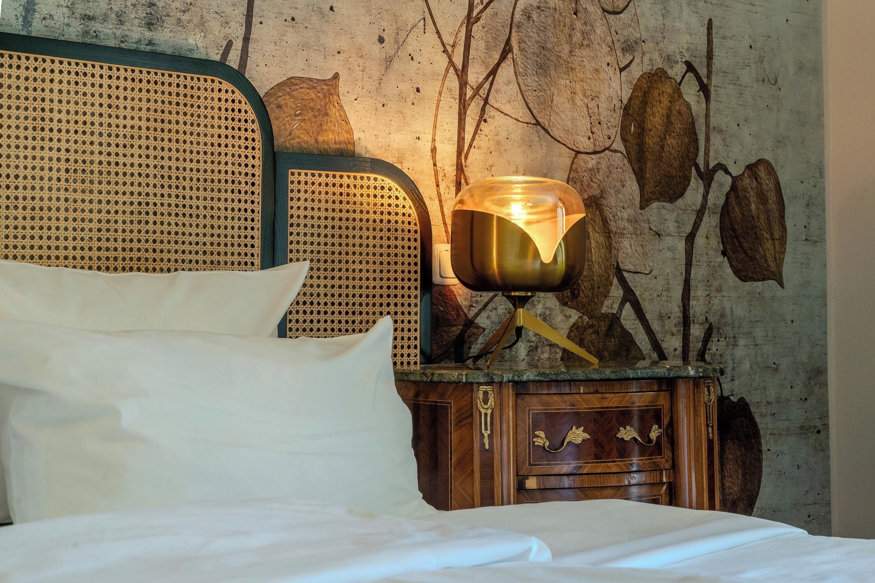 Una cama bien hecha con sábanas y almohadas blancas, una mesita de noche de madera con una lámpara, y una pared con un diseño rústico que incluye ramas y hojas.