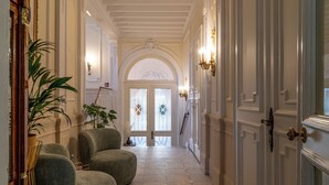 Un pasillo con piso blanco, dos sillones verdes, una planta en maceta y una puerta con picaporte de latón.