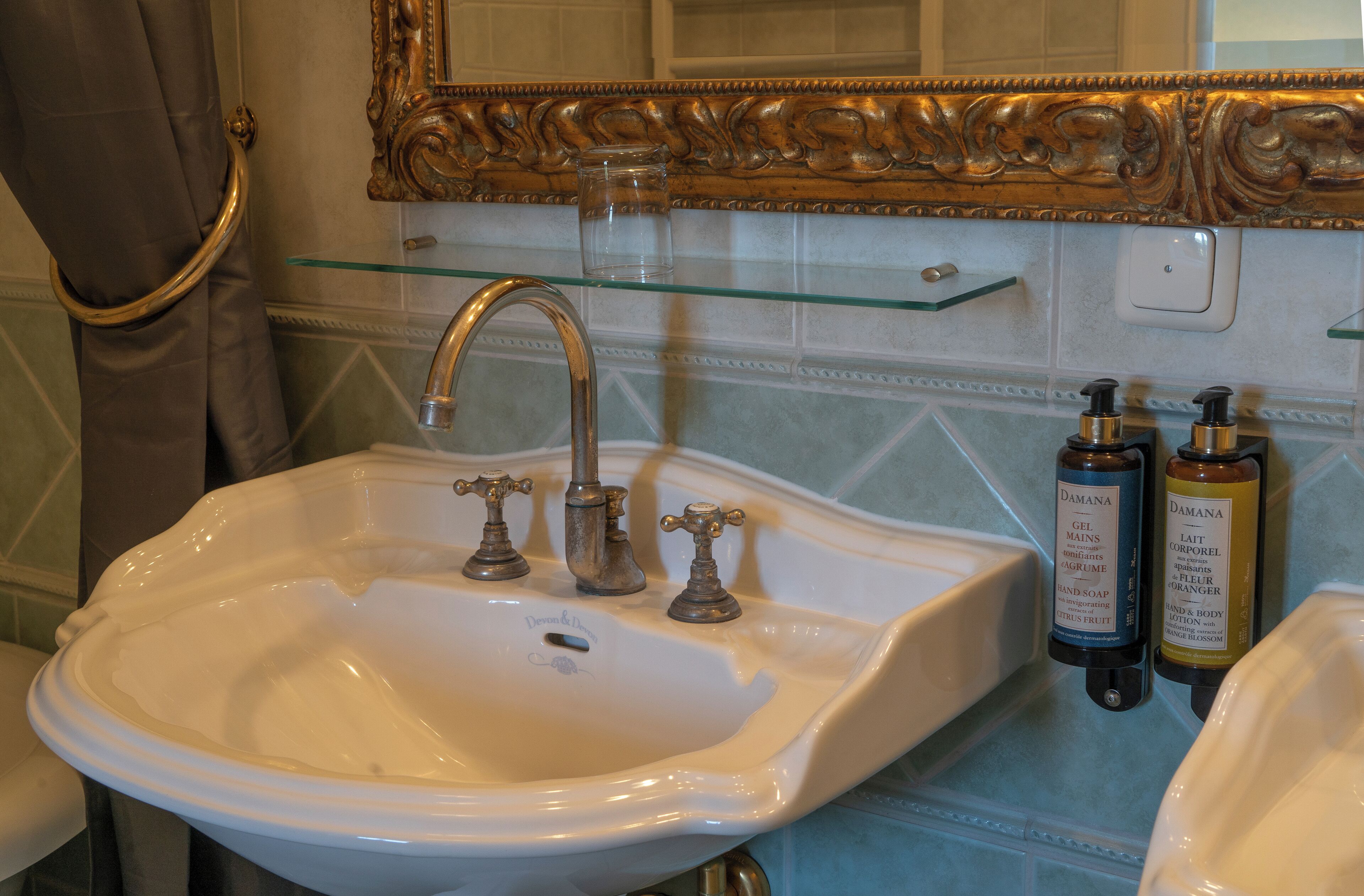 A bathroom with a white sink, two faucets, a glass shelf above the sink, and two bottles of lotion on the wall.