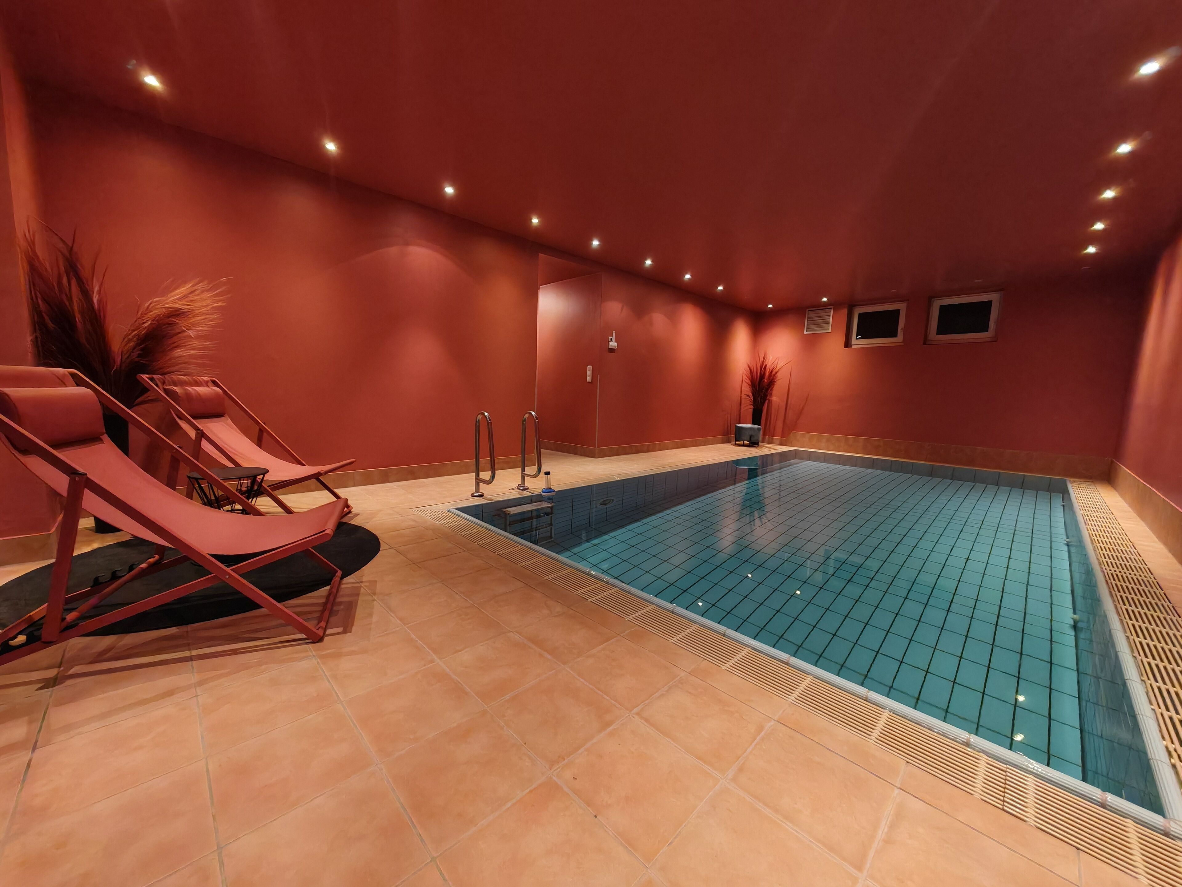 A swimming pool with lounge chairs and a small table, surrounded by red walls and recessed lighting.